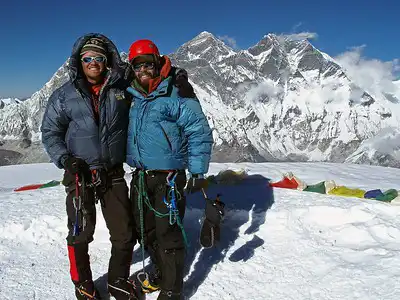 Two climbers standing on the summit of Ama Dablam with Everest and other Himalayan peaks in the background.