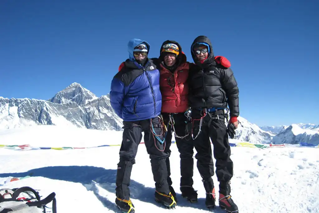 Three climbers standing on the summit of Ama Dablam with snow-covered peaks in the background.