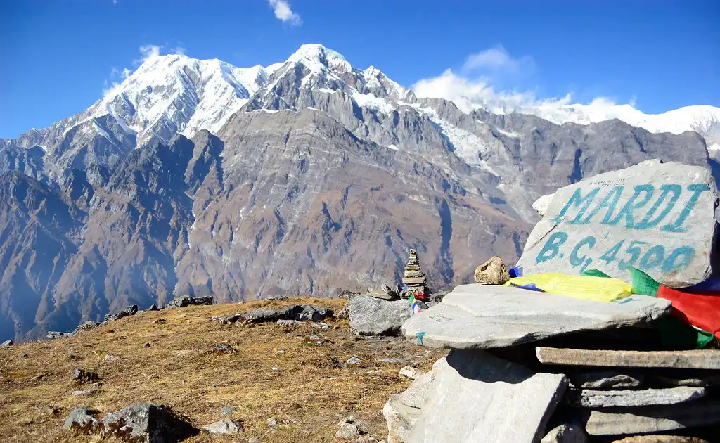 A stone at Mardi Himal Base Camp marked "MARDI B.C. 4500" with colorful prayer flags, set against a backdrop of towering snow-capped mountains.