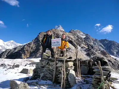 Two trekkers standing on a stone platform at Mardi Himal Base Camp, holding a sign that reads "Mardi Himal Base Camp 4500m," with snow-capped peaks in the background.