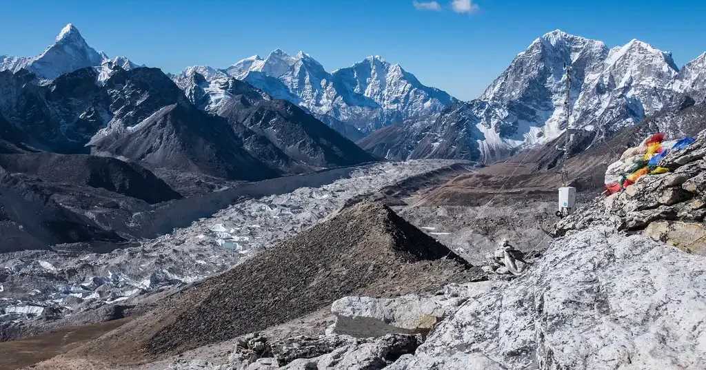 A panoramic view of the Himalayas from Kalapatthar, with rugged mountains, glaciers, and prayer flags visible in the foreground.