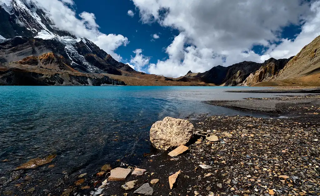 A stunning view of Tilicho Lake with clear turquoise water surrounded by rugged mountains and snow-covered peaks under a bright blue sky with scattered clouds.