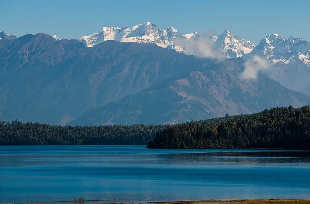 A serene view of Rara Lake in Mugu, Nepal, with clear blue waters reflecting the dense forest and the majestic snow-capped mountains in the background under a clear sky.