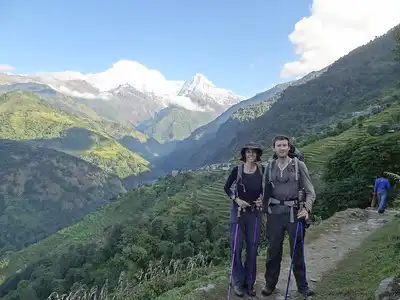 Two trekkers standing on a trail with trekking poles, with snow-capped mountains and terraced green hills in the background during the ABC trek.