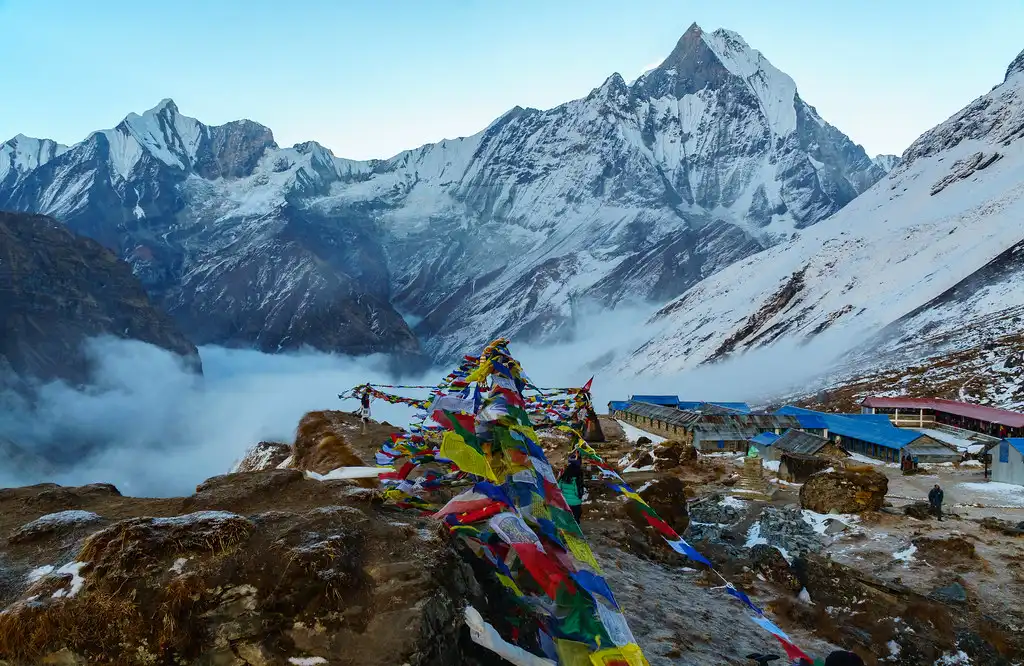 Prayer flags flutter in the wind at Annapurna Base Camp, with the snow-capped peak of Machhapuchhre in the background and a blanket of clouds below.