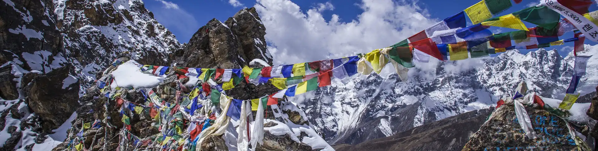 Colorful prayer flags strung across a rocky mountain pass with snow-capped peaks in the background, signifying the Renjo La Pass in the Himalayas.