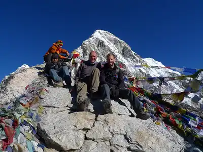 A group of trekkers sitting on rocks at the summit of Kala Patthar, surrounded by colorful prayer flags with a snowy peak in the background.