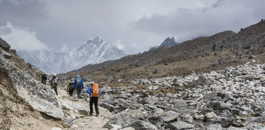 Trekkers walking on a rocky trail from Dingboche to Lobuche, with snow-capped mountains visible in the distance under a cloudy sky.