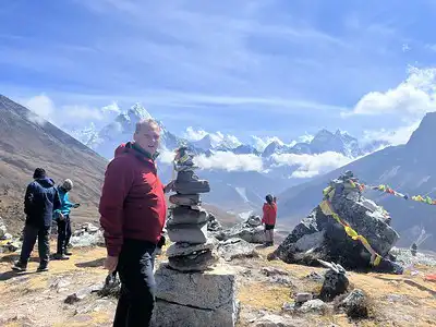 A hiker stands beside a rock cairn on a trail with a breathtaking view of the Everest mountain range in the background, surrounded by other trekkers.