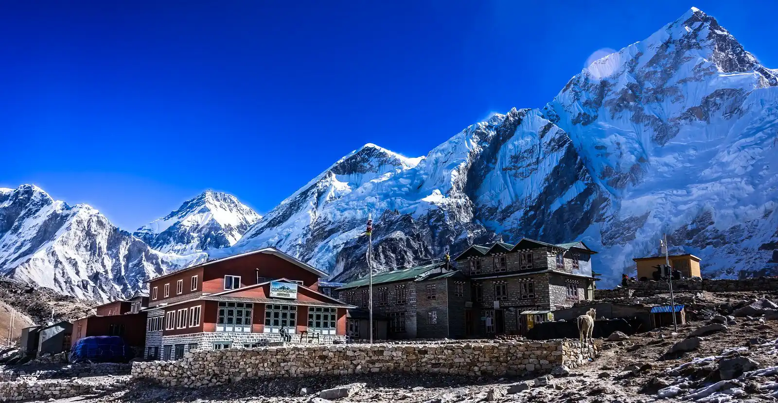 A view of Gorakshep, the last small settlement before Everest Base Camp, with its buildings set against the backdrop of towering, snow-covered Himalayan peaks under a vibrant blue sky.