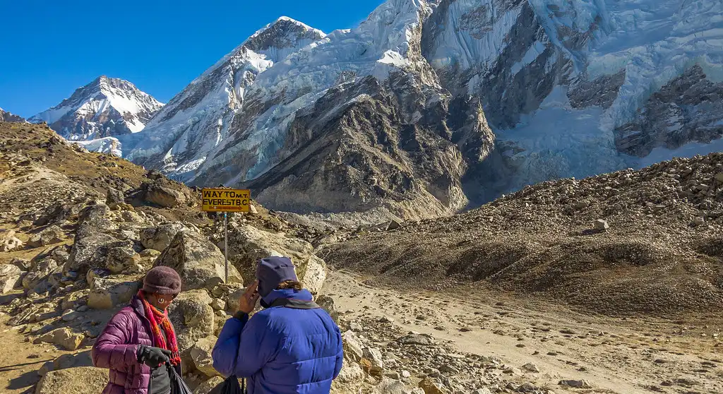 Trekkers on the rocky path from Gorakshep to Everest Base Camp, with a sign indicating the way and the towering, snow-covered Himalayan peaks in the background.