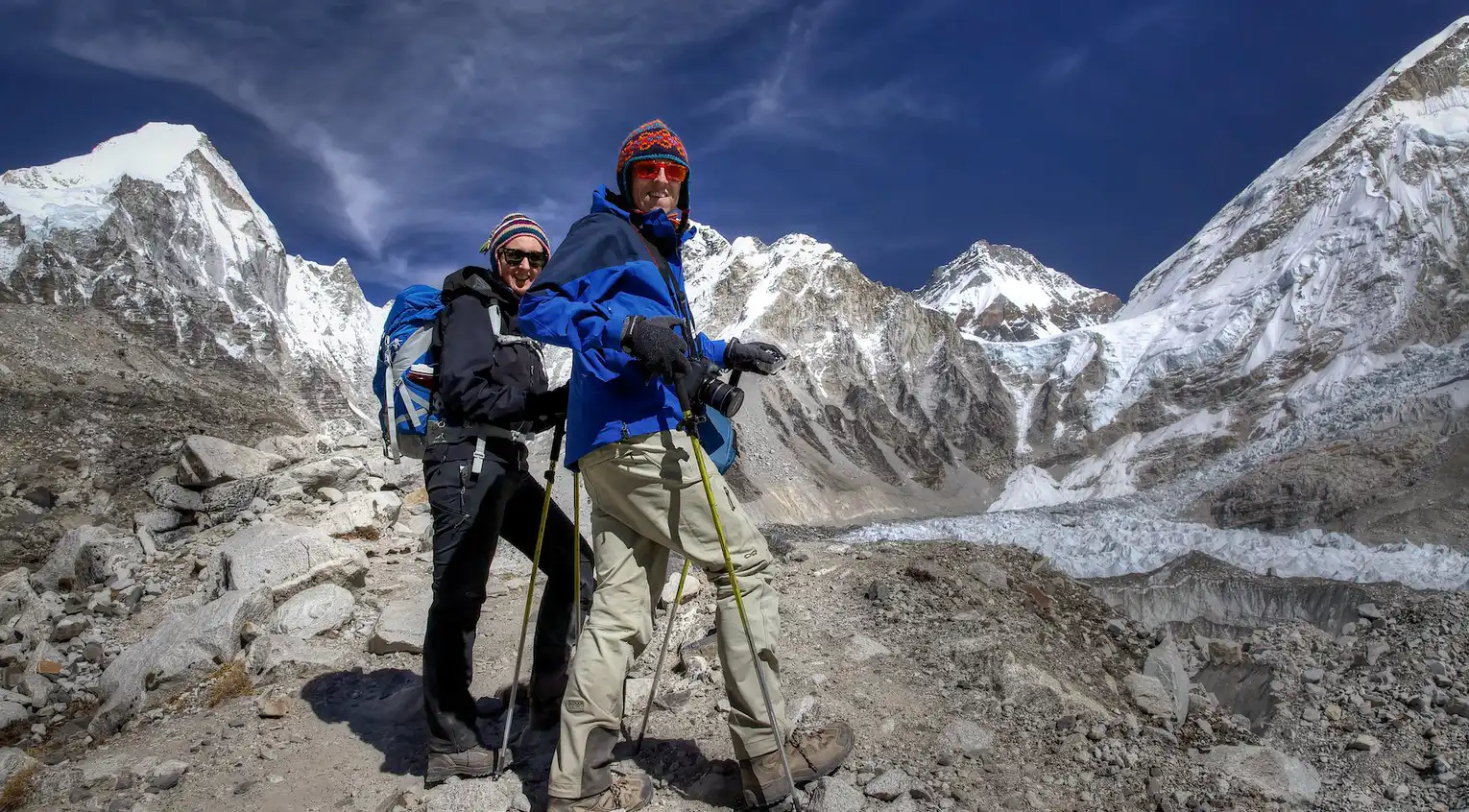 Two trekkers pose with trekking poles on a rocky trail between Gorakshep and Everest Base Camp, surrounded by towering snow-covered mountains and glaciers.