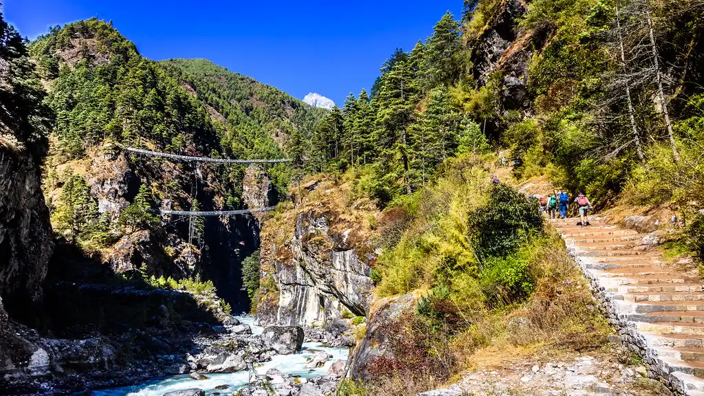 Trekkers crossing the Hillary Suspension Bridge over a river in the Everest region, surrounded by lush forests and steep cliffs under a clear blue sky