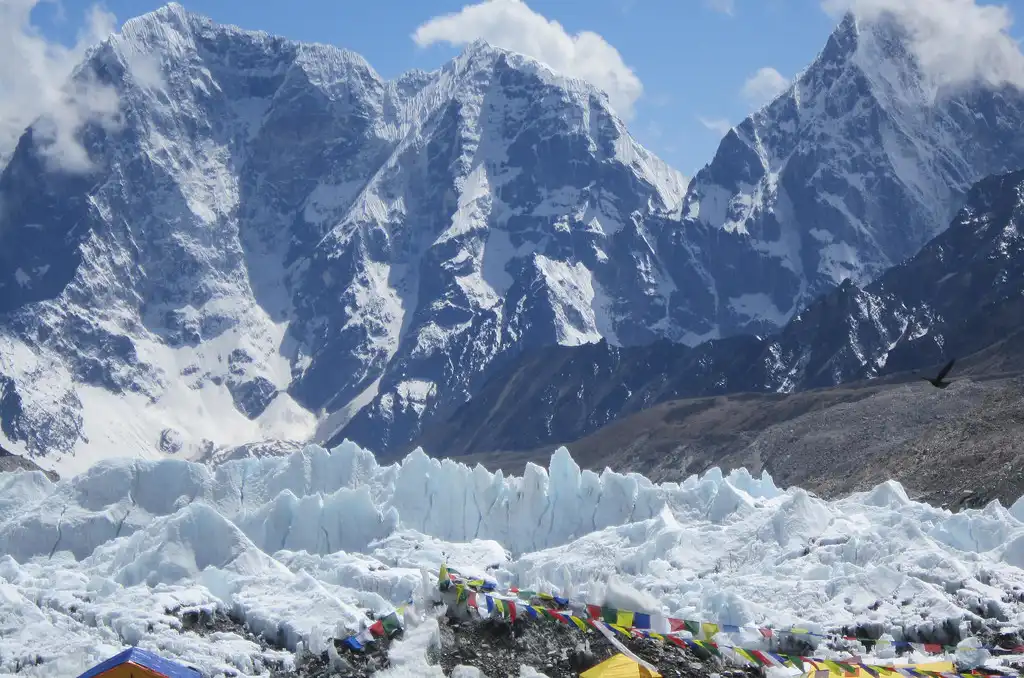 A view of the Khumbu Glacier with towering Himalayan peaks in the background, colorful prayer flags, and tents in the foreground.