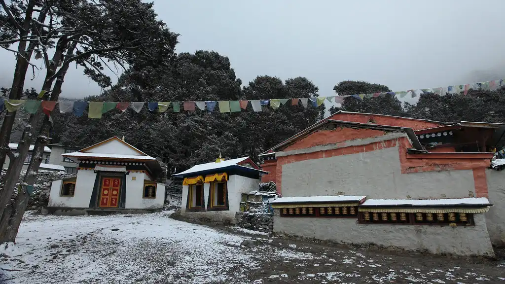 Khumjung Monastery buildings with prayer wheels and prayer flags in a snow-dusted courtyard, surrounded by trees.