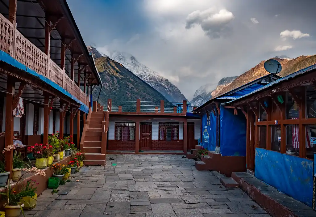 A traditional lodge at Chhomrong, Nepal, with stone-paved courtyards, vibrant flowerpots, and a backdrop of misty mountains under a cloudy sky.