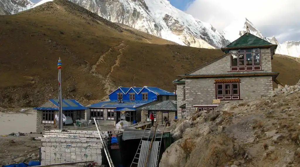 A lodge in Gorakshep, featuring traditional stone architecture with blue roofs, set against a backdrop of barren hills and snow-covered Himalayan peaks.