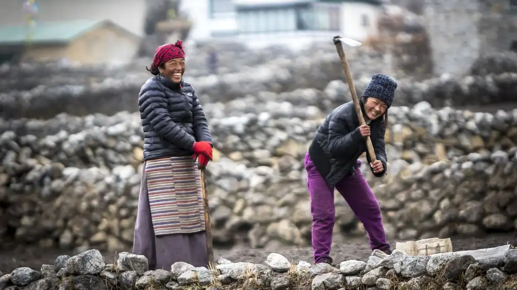 Two women from Khumjung village in Nepal working outdoors, one smiling while the other uses a tool to break the ground.