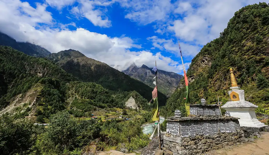 A scenic view of Phakding village in the Himalayas, featuring a traditional stupa with prayer flags, and a lush, green valley surrounded by tall mountains under a blue sky