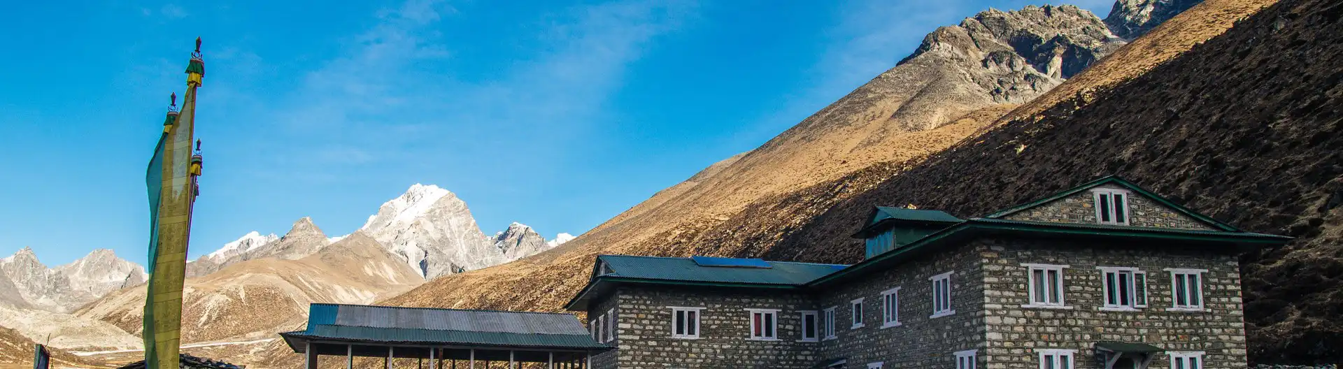 A stone building in Pheriche, Nepal, surrounded by towering Himalayan mountains under a clear blue sky, with traditional prayer flags in the foreground.