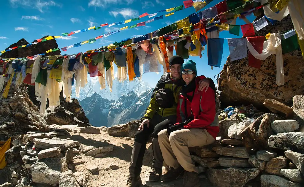 A couple embracing at Renjo La Pass, adorned with colorful prayer flags, with a clear view of snow-capped mountains in the background.