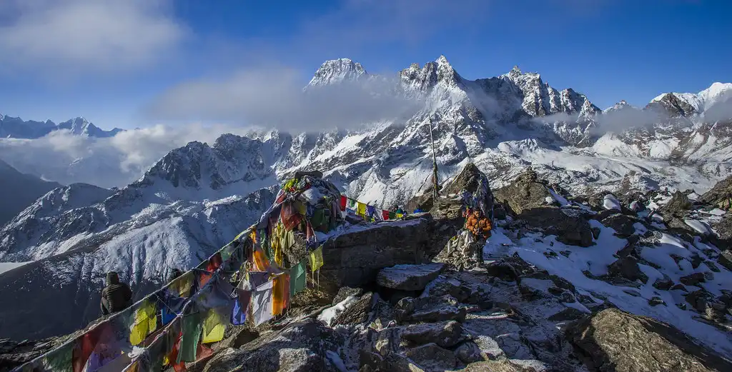 Hikers at the summit of Gokyo Ri surrounded by colorful prayer flags, with panoramic views of snow-covered Himalayan peaks under a clear blue sky.