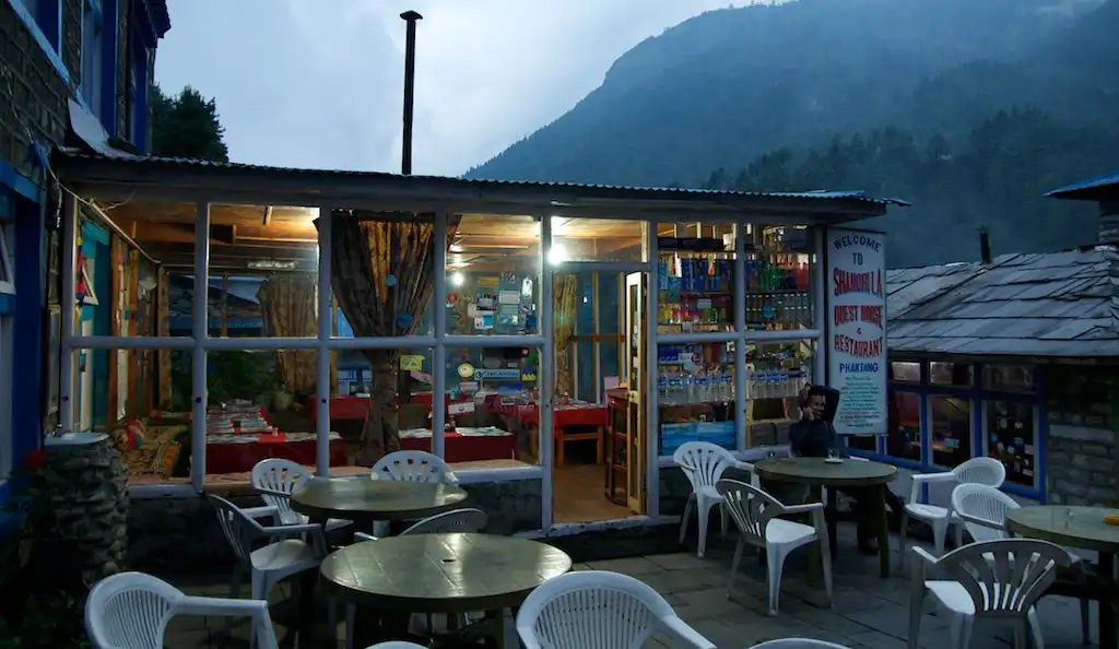 A cozy tea house in the village of Phakding, Nepal, with outdoor seating and a welcoming atmosphere during dusk, set against a backdrop of misty mountains.