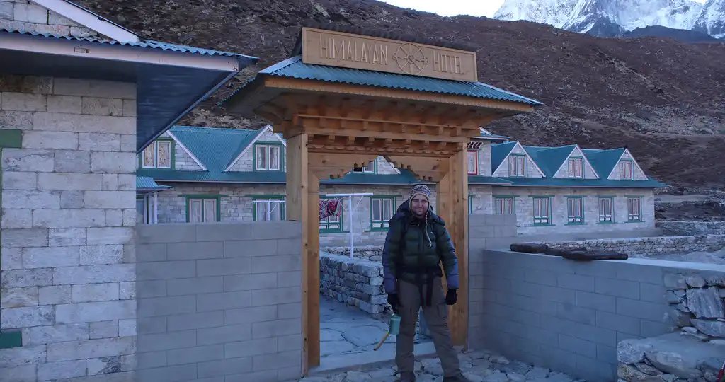 A trekker standing in front of the entrance to a lodge in Pheriche, Nepal, with stone buildings and snow-capped mountains in the background.