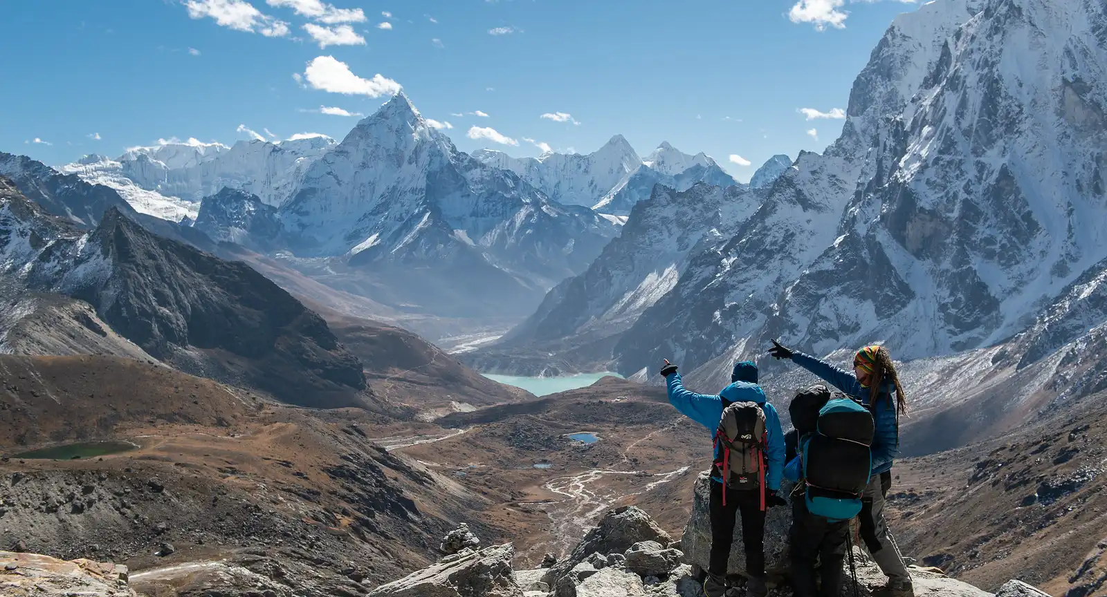 Group of trekkers at Chola Pass pointing towards a distant turquoise lake surrounded by rugged mountains and snow-capped peaks under a clear blue sky.