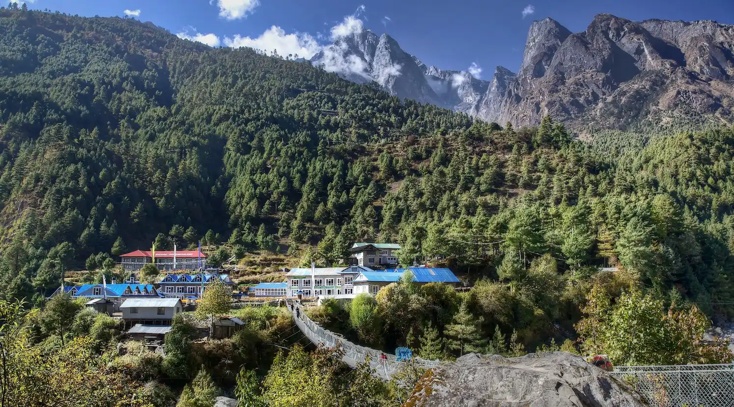 A picturesque view of a settlement in Phakding, nestled amidst lush green pine forests with towering rocky mountains and drifting clouds in the background.