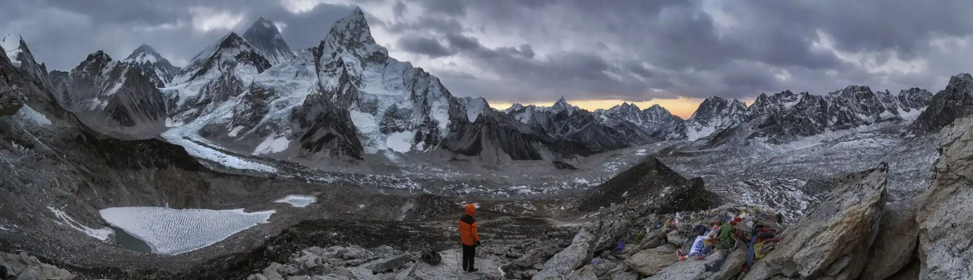 A trekker stands on the rocky summit of Kala Patthar, surrounded by snow-covered Himalayan peaks and glaciers, with Mount Everest visible in the background.