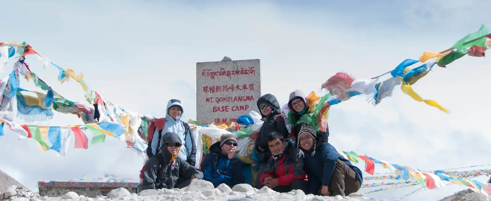 A group of trekkers poses for a photo at the North Everest Base Camp, with prayer flags draped around a sign marking the camp's location.