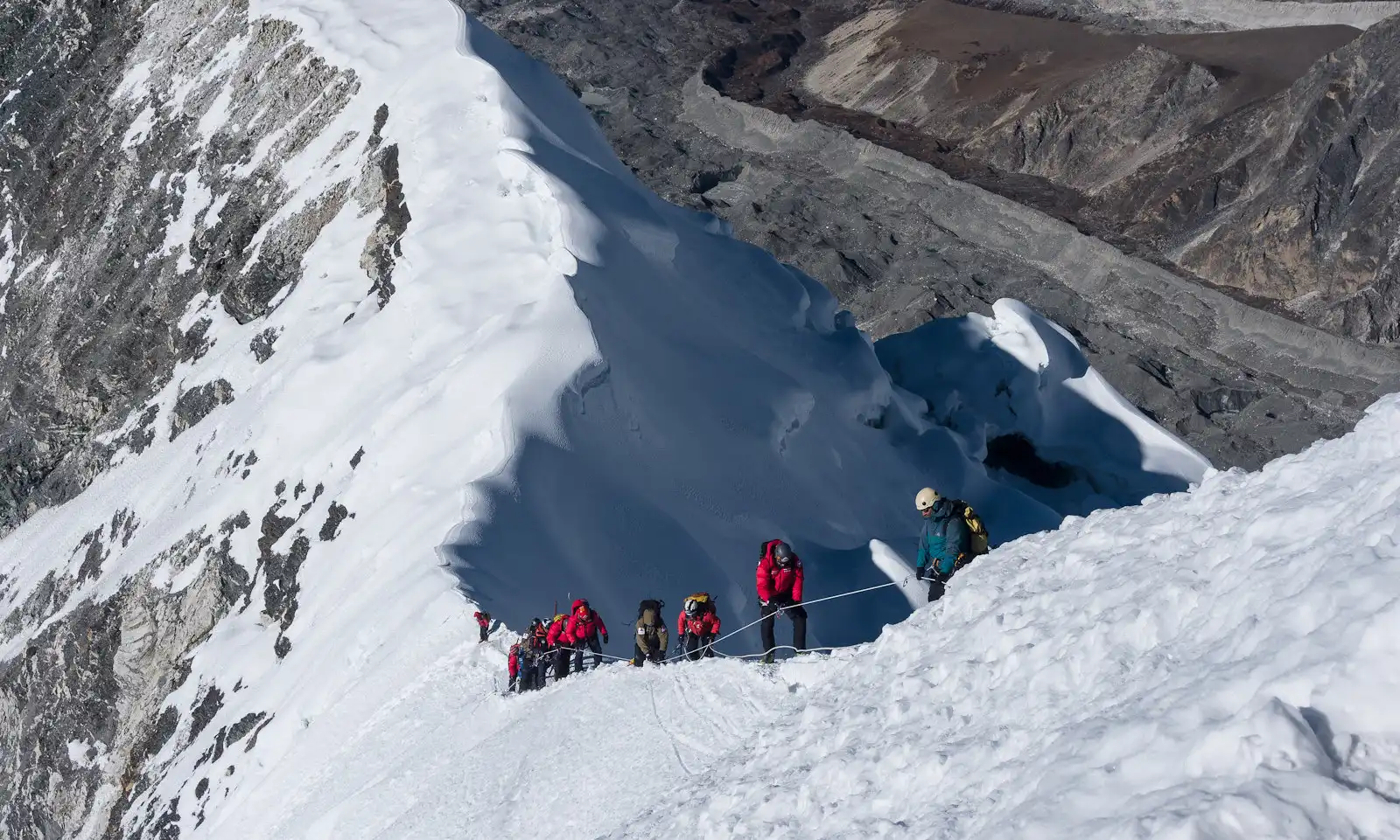 A group of climbers ascending the snow-covered ridge of Island Peak, navigating a challenging route with safety ropes, against a backdrop of rocky and icy terrain.