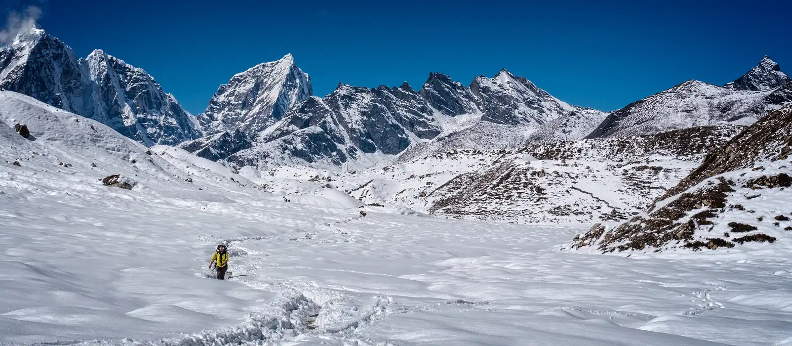 A climber advancing through a snow-covered landscape with towering, jagged mountains in the background under a clear blue sky.