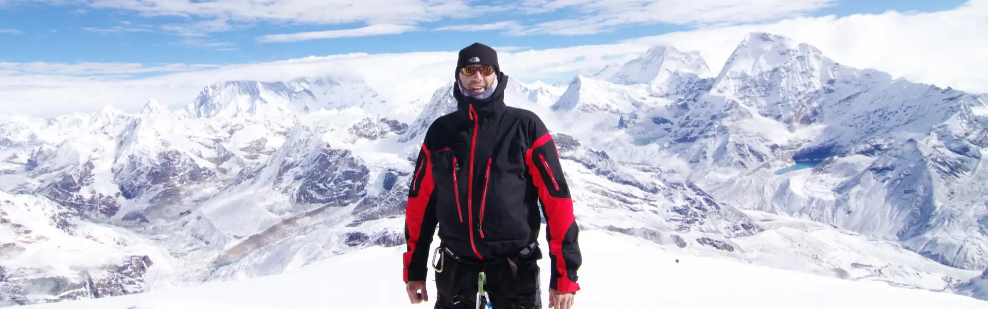 Man standing on the summit of Mera Peak with panoramic view of snowy Himalayan mountains in the background.