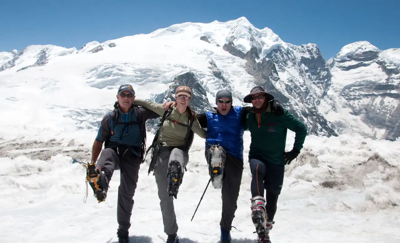 A group of climbers smiling and posing with one leg lifted, standing on snow-covered terrain, with a backdrop of towering snow-capped mountains during their ascent to Mera Peak.