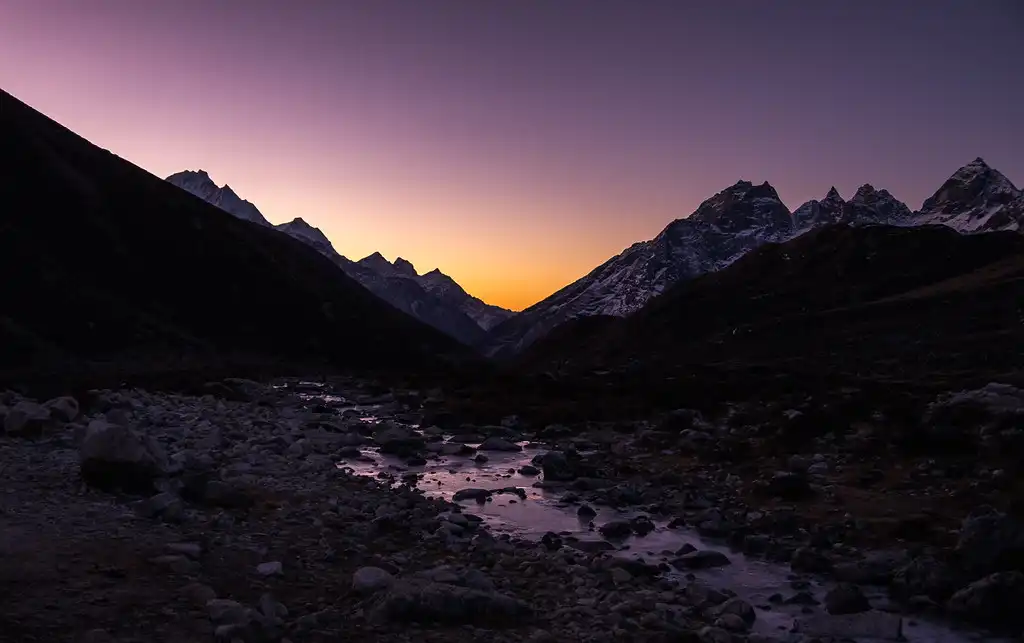 A peaceful early morning scene at Thangnak, with a stream flowing through rocky terrain, silhouetted mountains in the background, and a glowing orange and purple sky at dawn.