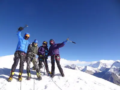 Group of climbers celebrating on a snowy peak with ice axes and harnesses, with clear blue skies and Himalayan mountains in the background.