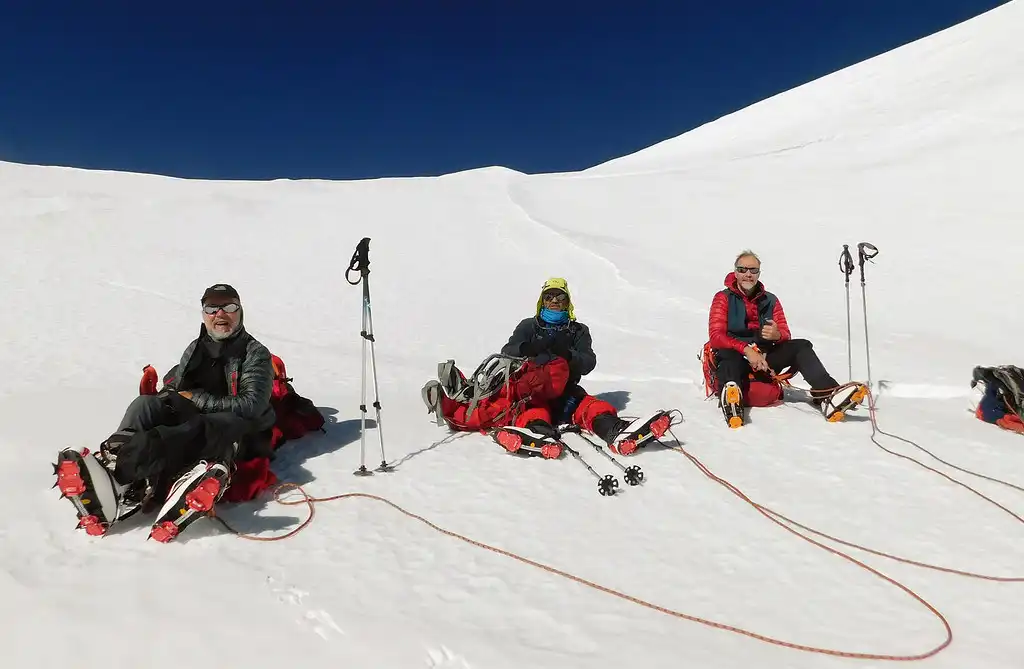 Three climbers resting on the snowy slopes of Mera Peak, sitting in the snow with their gear and trekking poles.