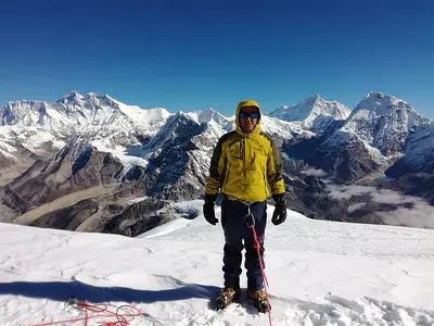 A climber standing on the snowy summit of Mera Peak, with the stunning Himalayan mountain range in the background under a clear blue sky.